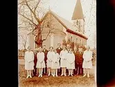 Konfirmander, 7 flickor, 5 pojkar och kyrkoherde Berntsson.
Norrbyås kyrka i bakgrunden.