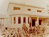 Tvåvånings villa med balkong, burspråk och veranda. Elva byggnadsarbetare framför huset.
Byggmästare Gothard Gustafsson