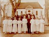 Konfirmander, 8 flickor, 5 pojkar och prästen.
Ödeby kyrka i bakgrunden.