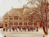 Trädgårdsskolan, tvåvånings skolbyggnad i tegel, skolbarn med lärarinna på skolgården.
Fröken Sofia Andersson