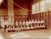 23 gymnaster i gymnastikhallen.
Gymnastiklärarinnan fröken M. Hallström