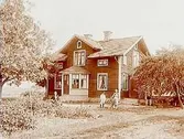 En och en halvplans bostadshus med frontespis och glasad veranda.
En vuxen och två pojkar framför huset.
Änkefru Anna Lovisa Olsson