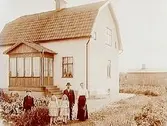 En och en halvplans bostadshus med brutet tak och glasad veranda. 
Familj 5 personer framför huset.
Per Hansson