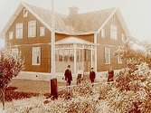 Vinkelbyggt en och en halvvånings bostadshus med glasad veranda.4 personer framför huset.August Johansson.