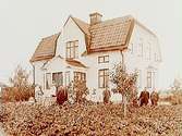 En och en halvplans villa med burspråk, glasad veranda, frontespis och brutet tak.
10 personer framför villan. 
P.G. Broström.