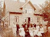 En och en halvvånings bostadshus med takhuv, 14 personer framför huset.G.F. Öhlin