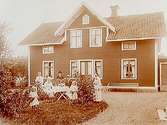 En och en halvvånings bostadshus med takhuv, separat köksingång.
7 personer vid kaffebordet framför huset.
Albin Karlsson