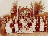En och en halvvånings bostadshus med takhuv och förstubro med snickarglädje.
Bröllop. Brudpar och bröllopsgäster framför huset.
Aug. Andersson