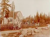 En och en halvvånings villa med brutet tak, vinkelbyggd, med förstebro. Vinkelbyggt uthus till höger om villan. 2 kvinnor och 3 barn framför uthuset. 2 män åkande motorcykel med sidovagn av märket Indian.
Sergeant G. Gustafsson