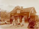 En och en halvvånings bostadshus med takhuv, förstebro och förstuga.
7 personer och en hund framför huset.
R. Bergström