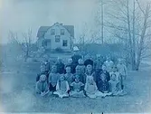 21 skolbarn med fröken Mathilda Löthgren. 
Byggnaden i bakgrunden kan vara klockargården Tysslinge. Mathilda Löthgren var lärare i Tysslinge kyrkskola.