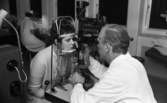 Kerstins Ögonoperation, Åtta poliser 31 okt 1967

Läkare undersöker ena ögat på en patient med en apparat. I bakgrunden sitter en sjuksköterska och skriver.