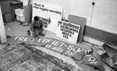 1 maj förberedelser 26 april 1967 

En man står på knäna och målar med en pensel på en banderoll inför 1 maj. Bakom honom står det skyltar lutande mot ett element.