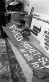 1 maj förberedelser 26 april 1967

Två män håller i en banderoll och förbereder inför 1 maj. Bakom dem står det några plakat mot väggen.