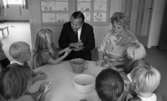 Barndaghem 9 september 1966

Vid ett bord sitter en förskollärare och en man i  svart kostym med barnen och leker med trolldeg. Bakom dem ser man teckningar på väggen.