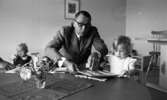 Barndaghem 9 september 1966

Vid ett bord hjälper en man i grå kostym en flicka med ett pussel. På bordet ligger det en duk och ett glas med blommor.