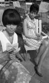 Systrarna Rynning igen, 7 juli 1965

Grythyttan utemöbler. Två kvinnor sitter utomhus på varsin stol. De är klädda i varsin sommarklänning. De har papper och pennor i sina händer. Kvinnan till vänster i förgrunden har en rosett vid barmen utanpå klänningen samt en fluffig, kort frisyr. Den andra kvinnan har en randig klänning samt kortklippt hår. Hon har pumps på fötterna.
