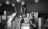 Drick vin gratis 31 juli 1965

Två män står i ett rum fyllt med lådor och kartonger. Tavlor står mot väggen i bakgrunden.