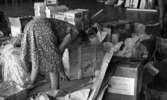 Drick vin gratis 31 juli 1965

Kvinna i klänning och glasögon står framåtlutad med vänster hand på golvet och med ryggen mot kameran. Runtomkring henne står fullt med pappkartonger och trälådor. På en pappkartong på golvet sitter en lapp med texten 
