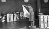 Drick vin gratis 31 juli 1965

Kvinna i klänning med glasögon rullar ihop karta med italiensk text. I bakgrunden syns tavlor med bl.a. antika motiv. En tavla föreställer en grekisk gud. Lådor står på golvet till höger på bilden.