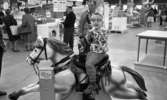 Stormarknaden, 31 juli 1965

Inne i Domus stormarknad. I förgrunden sitter en liten pojke på en automatisk leksakshäst med ett leksaksgevär i händerna. Åkturen kostar 50 öre vilket står bredvid på stolpen med myntintaget. I bakgrunden syns vuxna människor. Vissa skjuter kundvagnar framför sig. Varor till försäljning syns även som exempelvis cyklar, bord, tvättmaskin, frysbox etc.