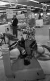 Stormarknaden, 31 juli 1965

Inne i Domus stormarknad. I förgrunden sitter en liten pojke på en automatisk leksakshäst med ett leksaksgevär i händerna. Åkturen kostar 50 öre vilket står bredvid på stolpen med myntintaget. I bakgrunden syns vuxna människor. Varor till försäljning syns även som exempelvis cyklar, bord, tvättmaskin, frysbox etc.
