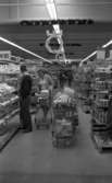 Stormarknaden, 31 juli 1965

I förgrunden står en man vid hyllan för skjortor till försäljning. En kvinna kommer gående för att ta sig förbi honom med en fylld kundvagn. Till höger om dem lite längre fram står två fyllda kundvagnar. Varor till försäljning kantar gången i affären såsom koppar, fat, kastruller etc. Ytterligare personer syns i bakgrunden.
