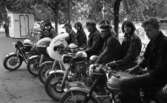 Knuttar och spättor 22 juni 1965.Tio stycken ungdomar med motorcyklar i förgrunden. Nyfiken kvinna kikar fram bakom glasskiosken.