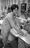 Damfrisörskor 8 juli 1965.

I förgrunden poserar en damfrisörska vid ett handfat inne på frisersalongen. I spegeln i bakgrunden syns en herre bli klippt av en annan frisörska.