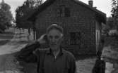 Hasselfors bruk 19 augusti 1965

En bruksarbetare iklädd keps och skjorta står framför en tegelbyggnad på Hasselfors bruk. Till vänster syns en väg. I bakgrunden till höger syns ytterligare en man.