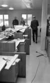 Handelsbanken Fellingsbro, 11 december 1965 

Fyra kostymklädda unga män inne på Handelsbanken i Fellingsbro. Träbitar, verktyg och papper ligger kringströdda på disken och skåpen. Lokalen och inredningen är under uppbyggnad.