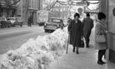 Halkiga pensionärer, 18 december 1965

En kvinnlig pensionär kommer gående på Drottninggatan en vinterdag.Hon är klädd i mörk lång kappa, pälsmössa med märke, mörka strumpor och skor på fötterna. I höger hand håller hon en käpp och i vänster hand en stor väska. Bredvid henne längs med trottoaren löper en stor, uppskottad snöhög. Framför henne står en kvinna och tittar på skyltningen i ett skyltfönster. Bakom henne går en herre som är på väg åt andra hållet. Framför honom ligger Stora Hotellet. En buss passerar och flera bilar står parkerade på gatan i bakgrunden. Ytterligare personer syns längre bort. Över gatan uppe i luften hänger juldekorationer.