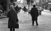 Halkiga pensionärer, 18 december 1965

I förgrunden på trottoaren kommer en äldre herre - en pensionär -  gående på Drottninggatan. Han har en käpp i handen. En kvinna kommer gående i motsatt riktning. Hon har ryggen mot kameran. Hennes hår är uppsatt. Till vänster syns skyltfönster. Längre fram på gatan i bakgrunden syns flera personer. På gatan står bilar parkerade. En buss passerar. Uppe i luften över gatan hänger juldekorationer.