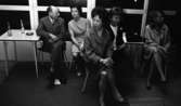 Minnenas 23 maj 1966

Fyra kvinnor och en man sitter på stolar invid en vägg med fönster. Vid mannen står ett bord med ett askfat med fimpar i samt två tomma Coca-Colaflaskor med sugrör i. Mannen har sin högra armbåge på bordet. Till höger syns ytterligare ett bord med en väska på.