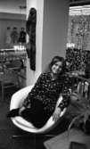 Modebilder 23 april 1966

En mannekäng klädd i blommig svart blus, svarta byxor och svarta skor sitter i en stol. Bakom henne syns fler stolar, en trappa, lampor samt ett antal skyltdockor i bakgrunden.