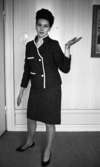 Modebilder 23 april 1966

En mannekäng klädd i kritstrecksrandig dräkt med kort kjol och svarta pumps håller vänstra handen utsträckt i en gest. På väggen syns en liten bit av en tavla som hänger där.