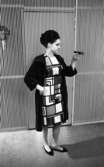 Modebilder 23 april 1966

En mannekäng med kort ljus klänning med mörka kvadrater på, svart kappa och svarta skor håller en konstgjord fågel i sin vänstra hand. Hennes hår är uppsatt. Bakom henne syns en randig vägg med randig dörr på samt en lampa på väggen.