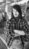 Modet 20 maj 1966

En ung man med långt hår klädd i rutig skjorta med mörk tröja inunder samt ljusa byxor står utomhus framför en sportaffär med sportartiklar i skyltfönstret.