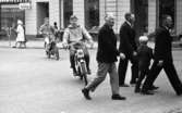 Mopedister 5 maj 1966

Två unga pojkar åker på mopeder på en gata i centrala Örebro. Framför dem passerar precis tre vuxna män och en liten pojke i mörka kläder. Tre damer syns i bakgrunden. De är klädda i kappor med hattar på huvudet. En barnvagn samt två parkeringsautomater syns i bakgrunden.