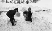 Snögrottor, 4 december 1965.

Två pojkar har skyfflat upp en hög med snö bakom staketet runt gräsmattan. Nu håller de på att gräva ur öppningen till grottan.