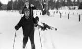 Hästnäs, 20 januari 1966

På bilden syns det tre ungdomar som är klädda i vinterkläder. Två pojkar står på skidor och flickan har ramlat och ligger på marken som är täckt med snö.