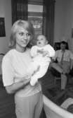 Mjölkande mammma 3 juli 1965

I förgrunden står en mor och håller sin baby i famnen. I bakgrunden sitter babyns far i en fåtölj.