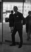 Andersson Gustavsson mode 23 april 1966

En manlig fotomodell visar herrkläder. Han är iklädd kostym, skjorta och slips. Han står vid en glasdörr och har högra handen på handtaget. Två kvinnor skymtar genom glasdörren samt glasväggen.