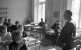 Engelsk lärare 6 april 1966

En lärare i engelska står framför eleverna i sin skolklass. De sitter ned i sina skolbänkar.















































































































or. Han går nedför en kort trappa utomhus.