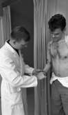 Ortopeden 28 februari 1966

En man i vit rock provar ut en armprotes på en man som står med bar överkropp. Bakom dem skymtar ett provrum.