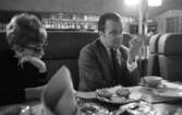 Gunnar Hellström, 21 februari 1966

Gunnar Hellström sitter tillsammans med en kvinna vid ett bord inne på en restaurang. Han håller upp sin vänstra hand i luften och har sin vänstra armbåge stödd mot bordet.  På bordet står ett fat med skorpor samt två askfat.  I bakgrunden skymtar en bardisk med flaskor.