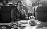 Gunnar Hellström, 21 februari 1966

Gunnar Hellström sitter vid ett bord inne på en restaurang och med en kopp te samt ett fat med skorpor framför sig. I bakgrunden skymtar en bardisk med flaskor samt två kvinnor ur restaurangpersonalen. Vid sidan om Gunnar skymtar en kvinna som man bara ser en liten del av.
