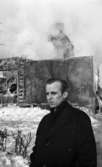 Nora-branden efteråt, 19 februari 1966

I förgrunden syns det en man som är klädd i en mörk rock. I bakgrunden skymtar det som är kvar av byggnaden efter brand. Istappar hänger på husfasaden.