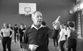 Mr Idla, 16 februari 1966

I förgrunden syns idrottsledare Ernst Idla. Han pekar med vänster pekfinger. Han är klädd i mörk tröja med vit krage. Han har en  scarf i halsen. I bakgrunden syns gymnaster. På väggen hänger en basketkorg. Till höger på bilden syns en väggklocka samt en stege.