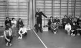 Mr Idla, 16 februari 1966

Mitt i bilden står idrottsledare Ernst Idla och instruerar åhörare och pekar med vänstra handen. Han är klädd i mörk tröja med vit krage, mörka byxor samt har en scarf i halsen. På ömse sidor om honom sitter flera gymnaster i en ring på golvet och lyssnar. På två långbänkar i bakgrunden sitter åhörare. Väskor står i mitten på golvet. På väggarna hänger ribbstolar.
